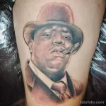 Фото тату: реалистичный портрет рэпера Notorious B.I.G. в шляпе и с сигарой, выполненный в черно-серых тонах на руке.