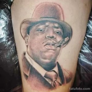 Фото тату: реалистичный портрет рэпера Notorious B.I.G. в шляпе и с сигарой, выполненный в черно-серых тонах на руке.