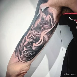 Фото тату в стиле blackwork с изображением волка и японской маски на руке мужчины.