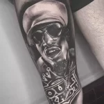 Фото тату 2Pac на руке в стиле реализм. Черно-белый портрет рэпера с солнцезащитными очками и надписью 'Thug Life'.