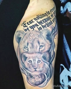 Фото тату львенка на плече с надписью 'Fear nothing in front of you because of God behind me'. Реалистичный рисунок татуировки в черно-серых тонах.