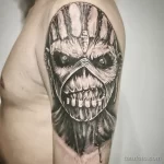 Фото тату: детализированный череп в стиле обложки альбома Iron Maiden, выполненный в черно-серых тонах на плече, с капающими кровью.