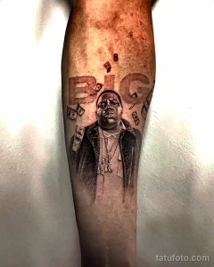 Фото тату на руке: реалистичный портрет The Notorious B.I.G. с изображением денежных купюр, выполненный в черно-серых тонах.