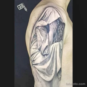 Фото тату: реалистичное изображение драпировки ткани, создающее эффект объема и глубины на плече. Татуировка выполнена в черно-серых тонах.