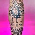 Фото тату Весы на икре, выполненное в технике blackwork. Изображение дерева с весами, символизирующее баланс и справедливость.