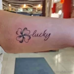 Фото тату: Черный контурный клевер с четырьмя листьями и надпись 'lucky' рядом с ним, нанесенные на руку. Элегантный и минималистичный дизайн.