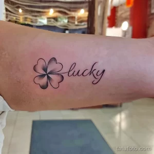 Фото тату: Черный контурный клевер с четырьмя листьями и надпись 'lucky' рядом с ним, нанесенные на руку. Элегантный и минималистичный дизайн.