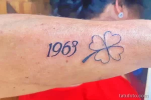 Фото тату: синий контурный рисунок четырехлистного клевера и цифр '1963' на запястье.