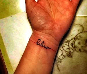 Фото тату: надпись 'believe' выполненная тонкой линией на запястье. Черно-белая татуировка, выглядит минималистично и вдохновляюще.