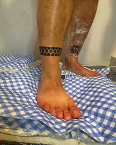 Фото тату: черный браслет в стиле трайбл, обхватывающий лодыжку, с геометрическим узором.