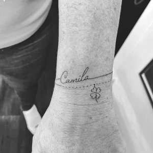 Фото тату на запястье: надпись "Camila" выполненная каллиграфическим шрифтом и рядом изображен четырехлистный клевер, выполненные в технике лайнарт.