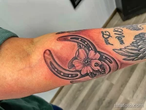 Черно-серое фото татуировки подковы с цветком сакуры и надписью 'Mom Dad' на предплечье.