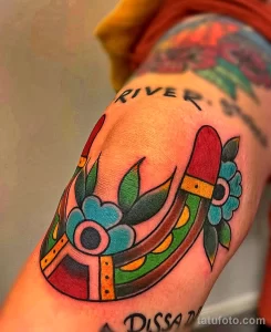 Фото тату подковы с цветком и надписью 'River' в традиционном стиле на руке. Цветной рисунок татуировки с четкими контурами и насыщенными цветами.