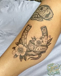 Фото тату подковы, украшенной розовыми цветами и лентой с надписью 'Mom'. Черно-серый рисунок татуировки на руке, выполненный в стиле традиционной татуировки.