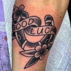 Фото тату подковы с надписью 'Good Luck', выполненной в традиционном стиле Old School на предплечье. Яркие цвета, четкие контуры, классический дизайн.
