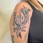 Фото тату подковы и цветов, выполненной в технике Blackwork на плече. Изящный рисунок, монохромная палитра, детализированные элементы. Прекрасный эскиз тату для ценителей минимализма.