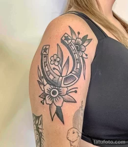 Фото тату подковы и цветов, выполненной в технике Blackwork на плече. Изящный рисунок, монохромная палитра, детализированные элементы. Прекрасный эскиз тату для ценителей минимализма.