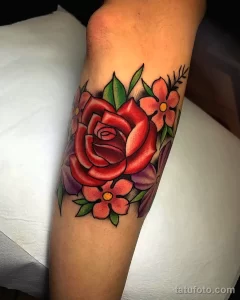 Фото тату розы на предплечье. Рисунок выполнен в классическом стиле с использованием красных, розовых и зеленых цветов. Эскиз татуировки изображает распустившуюся розу с листьями.