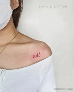 Фото тату: изящная надпись 'still' красным цветом, выполненная в технике лайнуорк на ключице девушки. Минималистичный и стильный рисунок.