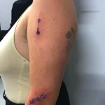 Фото тату с изображением веток с цветами, выполненных тонкими линиями в синих тонах на плече и ключице. Минималистичный стиль.