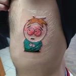 Фото татуировки с изображением Картмана из мультсериала South Park на бедре. Яркий, мультяшный стиль, выполненный в цвете.