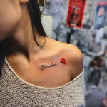 Фото тату: маленькая красная роза и надпись 'Elle rêve' на ключице девушки. Изящный рисунок татуировки в минималистичном стиле.