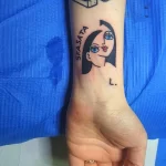 Фото минималистичной татуировки лица женщины с надписью "SFASATA" и точкой на предплечье. Черно-белое изображение в лаконичном стиле.