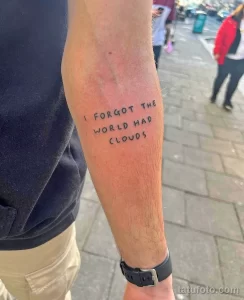 Фото тату с надписью 'I forgot the world had clouds' на предплечье, выполненной тонкой линией черным цветом. Минималистичный рисунок татуировки.