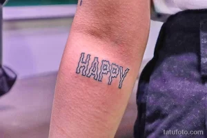 Фото тату с надписью 'HAPPY', выполненной в стиле ретро с использованием контура и заливки. Небольшой рисунок на руке, символизирующий позитив и радость.