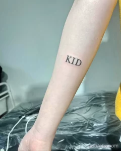 Фото тату: надпись 'KID' черным шрифтом на предплечье. Минималистичная татуировка, выполненная четкими линиями.