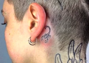 Фото тату: Небольшой рисунок скорпиона, выполненный в черном цвете, расположен за ухом на коротко стриженной голове. Минималистичный эскиз тату.