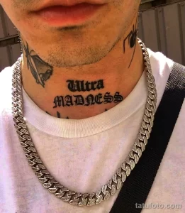 Фото тату: Надпись 'Ultra MADNESS' готическим шрифтом на шее мужчины. Черная татуировка с небольшим рисунком летящей птицы.