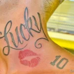 Фото тату надписи 'Lucky' с отпечатком губ и цифрой 10 на шее. Черно-серые оттенки, романтический стиль, уникальный дизайн. Подходит для тех, кто хочет увековечить важную дату или человека.