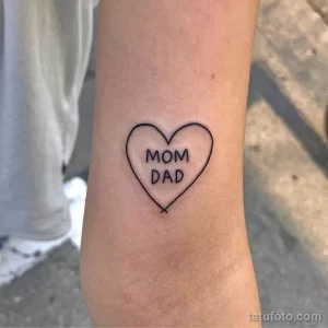 Фото тату на запястье: черное контурное сердце с надписью 'Mom Dad' внутри. Минималистичный рисунок татуировки для выражения любви к родителям.