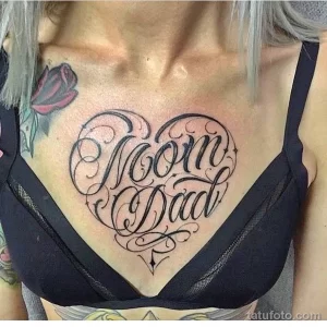 Фото татуировки в виде сердца с надписями 'Mom' и 'Dad' на груди. Черно-белый рисунок, каллиграфический шрифт, элегантный дизайн.
