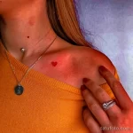 Фото тату: маленькая красная татуировка в форме сердца, расположенная на ключице, на фоне светлой кожи и одежды.