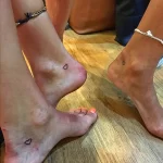 Фото тату: две крошечные черные татуировки в форме сердца, расположенные на лодыжках двух ног, на фоне деревянного пола.