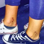Фото тату: две небольшие черные татуировки в виде сердца, расположенные на лодыжках обеих ног, на фоне джинсов и кед.