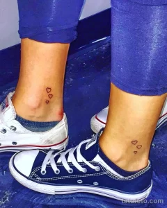 Фото тату: две небольшие черные татуировки в виде сердца, расположенные на лодыжках обеих ног, на фоне джинсов и кед.
