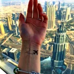 Фото тату: Татуировка на запястье с изображением самолета, летящего сквозь сердце, на фоне городского пейзажа, выполненная в черном цвете.
