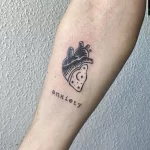 Фото тату: минималистичный рисунок сердца с луной и надписью 'anxiety', выполненный на предплечье. Эскиз татуировки, символизирующий борьбу с тревожностью.