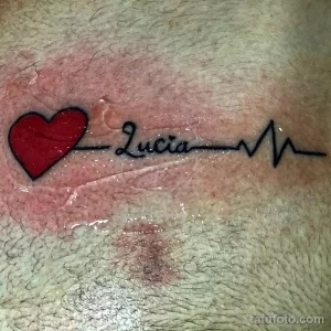 Фото минималистичной татуировки на коже, изображающей линию пульса с именем 'Lucia'. Простой и элегантный рисунок.