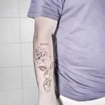 Фото тату на предплечье: реалистичная роза с листьями и римскими цифрами, символизирующими дату. Изящный рисунок татуировки в черно-серых тонах.