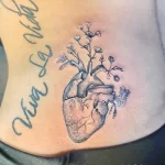 Фото тату сердца с растущими из него цветами и надписью 'Viva La Vida' на боку женщины. Черно-серый рисунок татуировки.