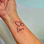 Фото тату надписи 'love' с изображением двух соединенных сердец на запястье. Минималистичный рисунок татуировки в черном цвете.