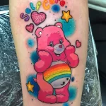 Фото тату розового Care Bear с радужным животиком, окруженного звездами и сердечками, на руке. Мультяшный рисунок татуировки выполнен в ярких и насыщенных цветах.