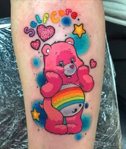 Фото тату розового Care Bear с радужным животиком, окруженного звездами и сердечками, на руке. Мультяшный рисунок татуировки выполнен в ярких и насыщенных цветах.