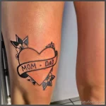 Фото тату на ноге: сердце с надписью "MOM & DAD", украшенное небольшими цветочными элементами. Черно-белый рисунок, символизирующий любовь к родителям.