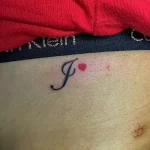 Фото тату на пояснице: буква 'J' и маленькое красное сердце, выполненные в тонком черном контуре.