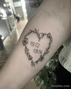 Фото тату на руке: сердце, обвитое цветами, с датами 1972 и 1976 внутри. Изящный рисунок татуировки в минималистичном стиле.